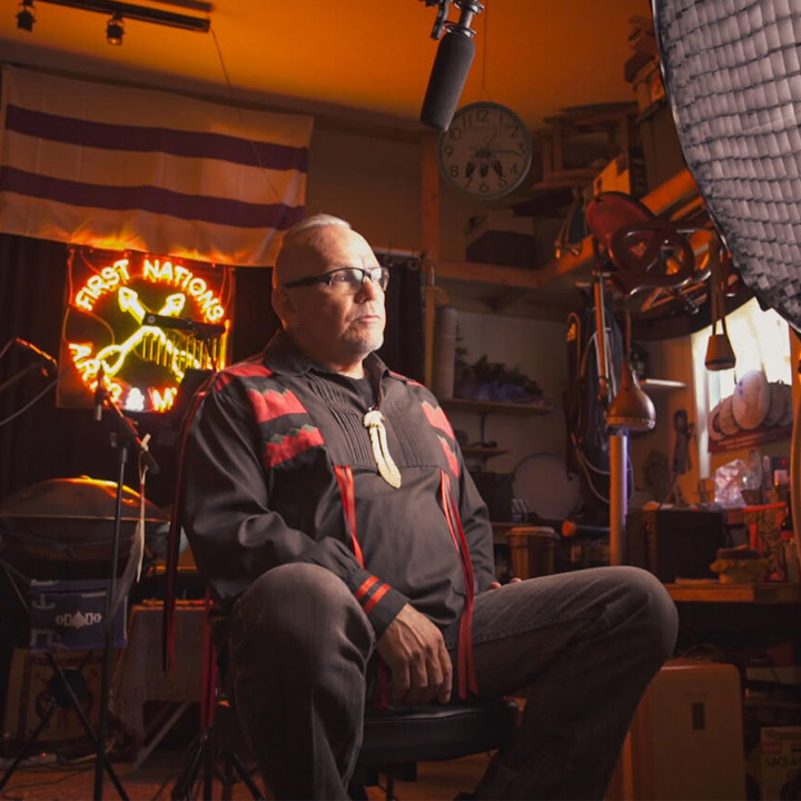World renown artist and musician David R. Maracle sits on a stool in a studio setting surrounded by lighting and recording equipment, including a large softbox light and an overhead microphone. Behind them, a neon sign reads “First Nations Art & Music”.