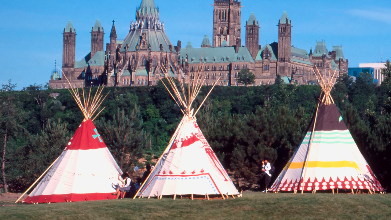 Three indigenous teepees stand on a grassy field in the foreground with Canada’s Parliament Hill in the background.