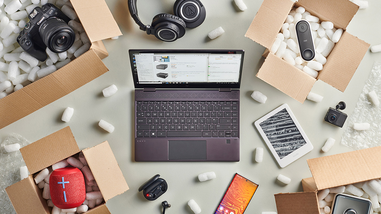 Top-down view of a laptop at the center surrounded by various tech items arranged in open cardboard shipping boxes. 