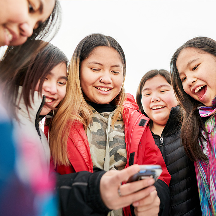 A group of children stand close together outdoors, looking down at a smartphone held by one child in the center. 