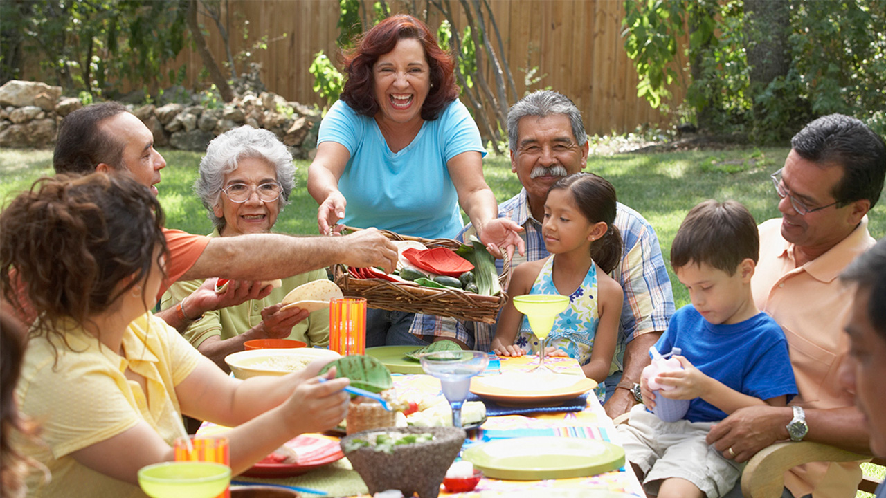 A multigenerational family sitting outdoors around a table, passing dishes and sharing a meal together.