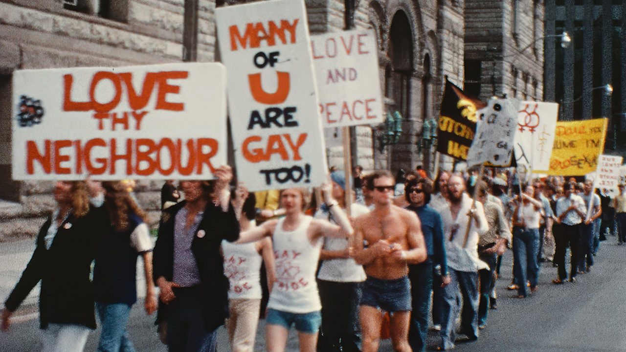 A group of people walking in a street parade holding signs with messages such as ‘Love thy neighbour,’ ‘Many of you are gay too!’ and ‘Love and peace.’