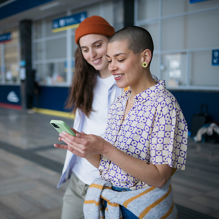 Two friends using smart phone while waiting for train at railway station.