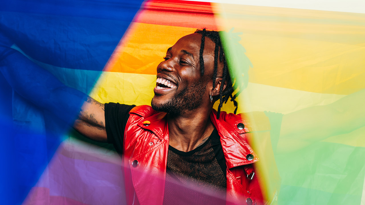 A smiling Black person wearing a red sleeveless jacket holds and waves a rainbow pride flag.