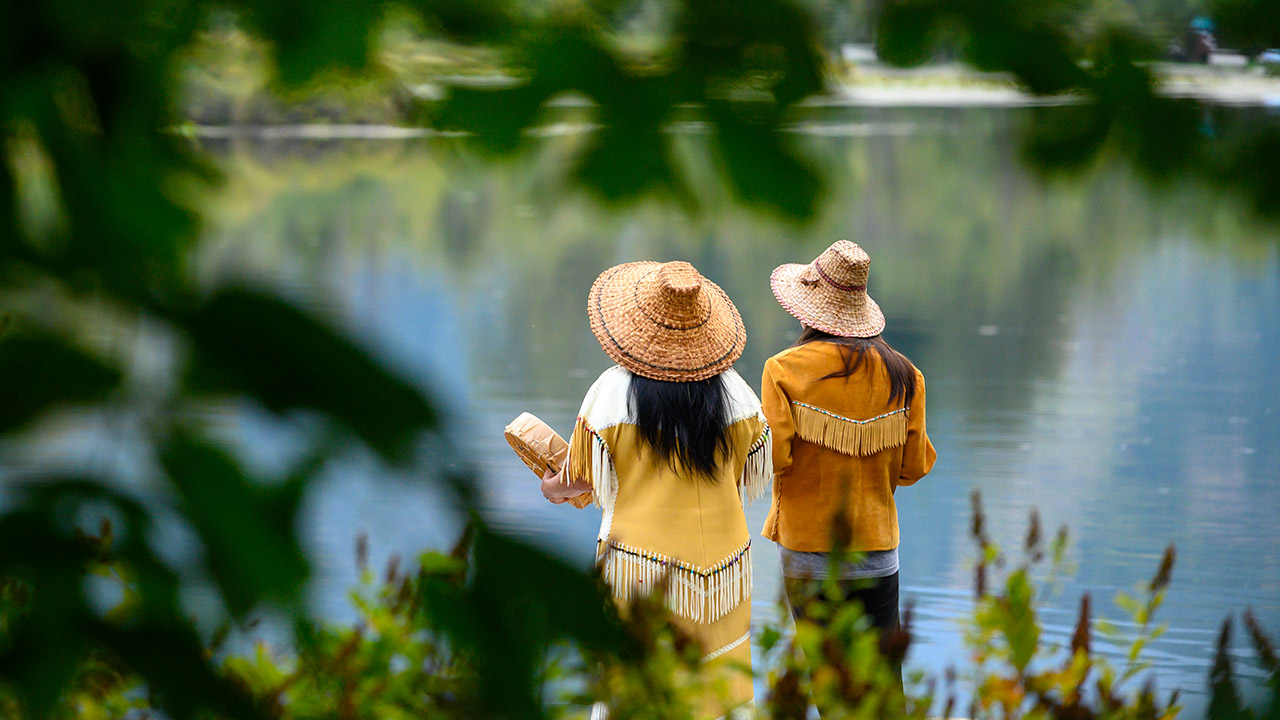 Two Indigenous people stand side by side by a calm lake, surrounded by lush green foliage.