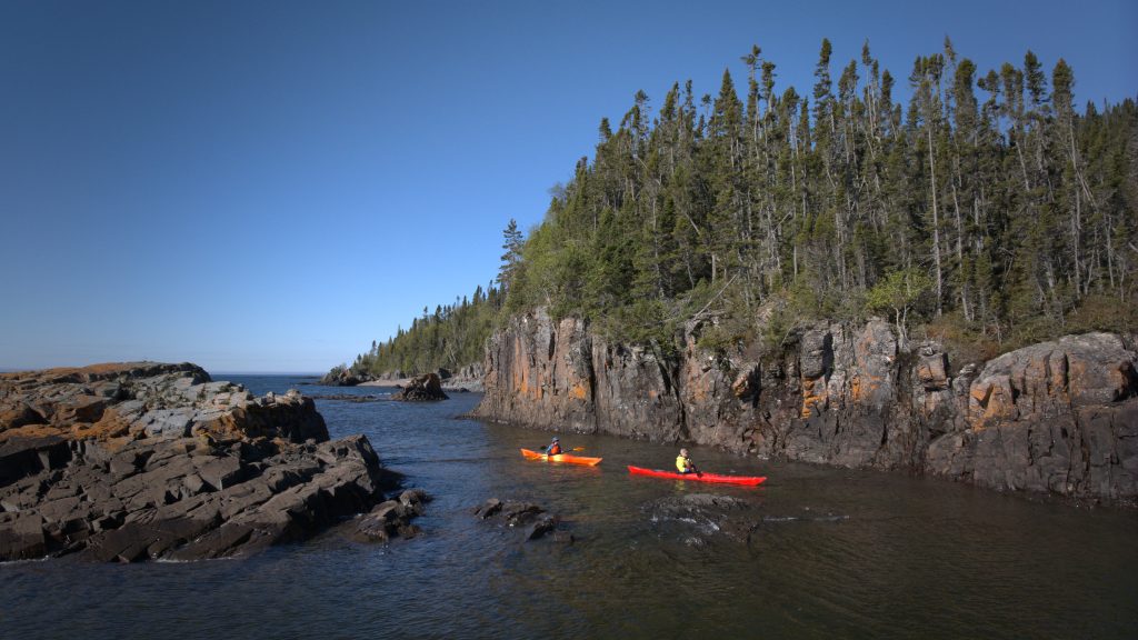 Two kayakers in bright orange and red kayaks paddle through a narrow, calm inlet bordered by rugged dark rock cliffs and dense evergreen forest beneath a clear blue sky.